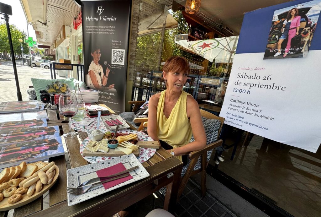 Helena Viñuelas sonríe sentada junto al cartel de la presentación de «El club de las que NO sudan» en Cattleya Vinos