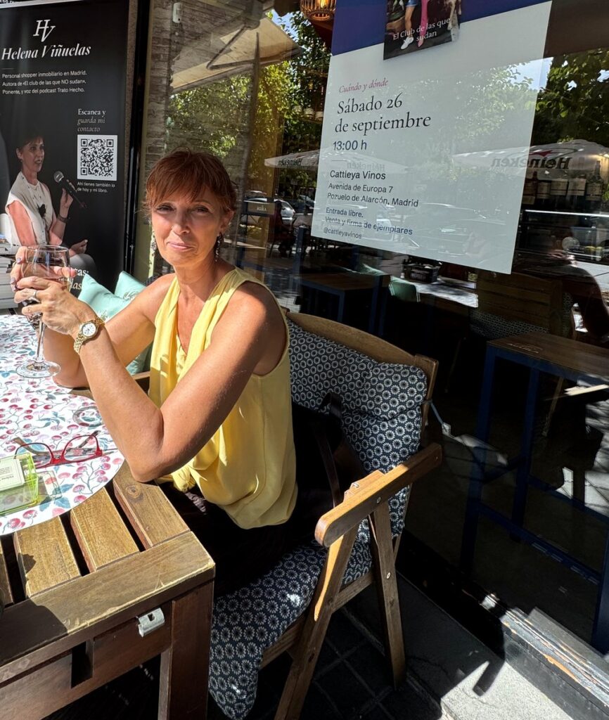 Helena Viñuelas, sentada en la terraza de Cattleya Vinos con una copa en la mano, delante del cartel que anuncia la presentación de su novela