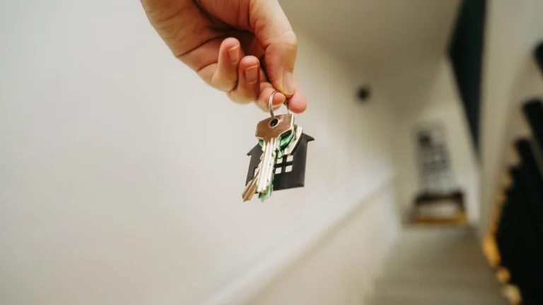 Mano que sostiene unas llaves con un llavero negro en forma de casa, en el pasillo blanco de una vivienda